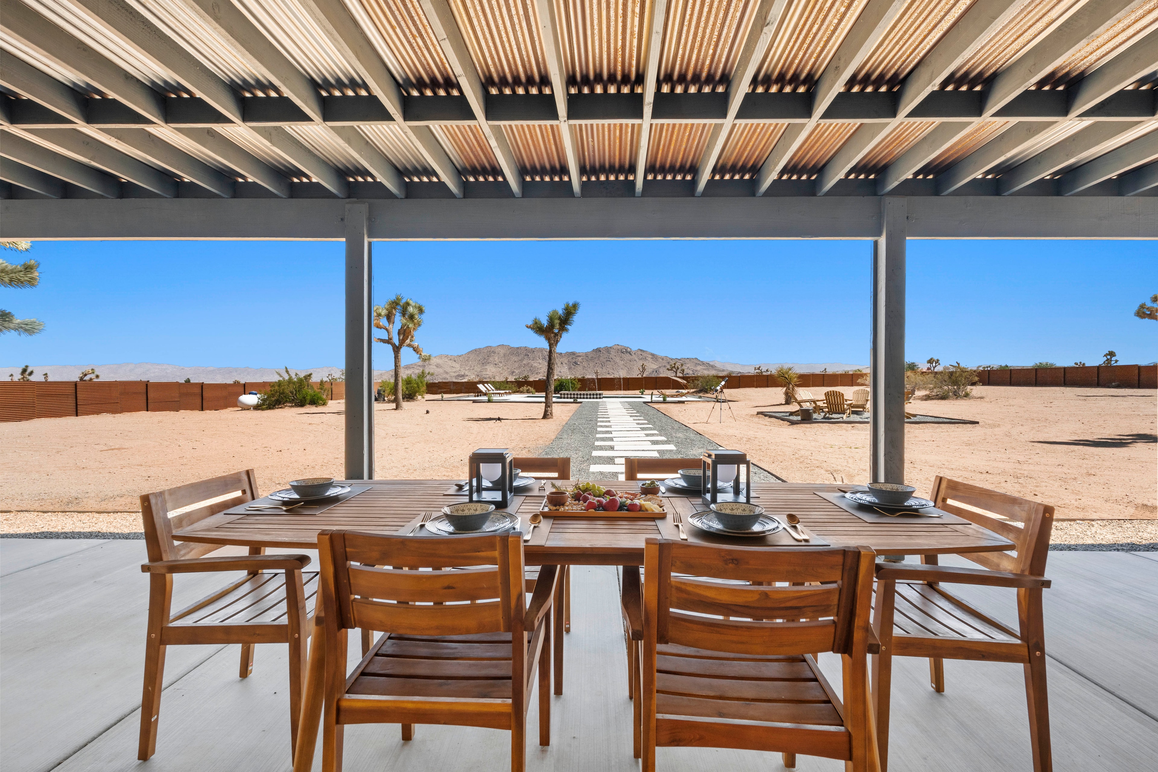 Covered outdoor dining patio with teak table set for six, looking out over the fenced desert yard with Joshua trees and mountain views